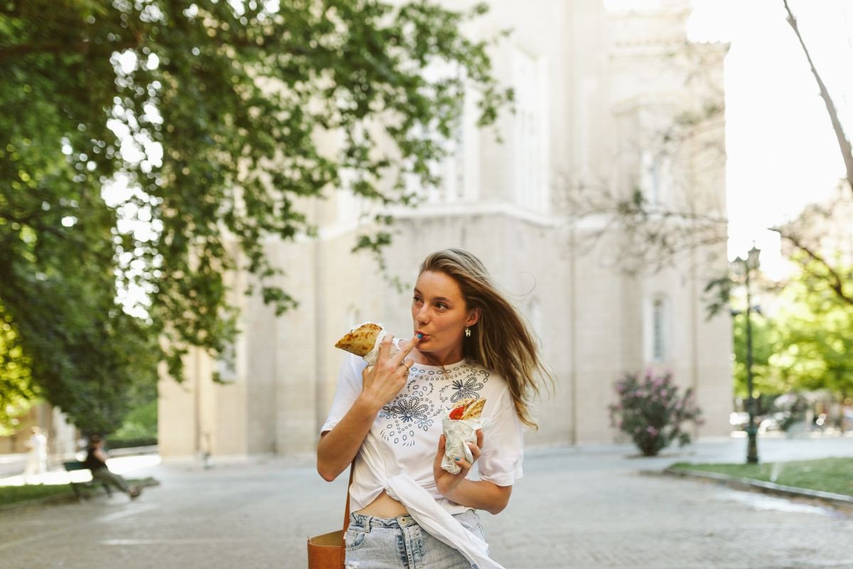 A woman eating Greek soubvlaki on a tree-lined street with a backdrop of a bright, historic building.